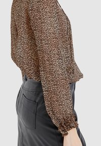 Blouse marron transparente avec un imprimé léopard, des manches élastiques et une coupe ample, associée à une jupe en cuir noir avec des poches.