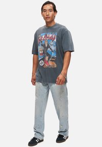 Grå grafisk t-shirt med en komisk stil som visar en karaktär klädd i svart, tillsammans med ljusa denimbyxor med färgfläckar.