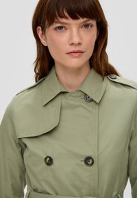 Jasnozielony płaszcz trenchowy z podwójnym zapięciem, dużymi guzikami i kołnierzem. Gładka tekstura, wcięta talia i epolety na ramionach.