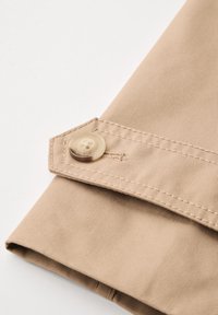 Beige-kangas, jossa on uritettu pinta, ja siinä on suuri, litteä nappi sekä ommeltu yksityiskohta taitetulla reunalla.