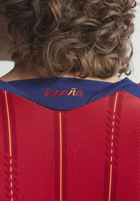 Osoba s kovrčavom kosom nosi sportsku majicu u crvenoj i tamnoplavoj boji s žutim prugama i natpisom "España" izvezenim na zadnjem ovratniku.