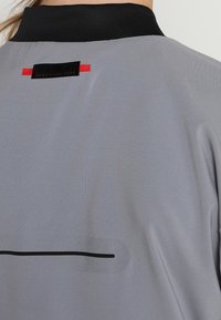 Grå jacka med svart krage, med en svart och röd etikett vid nacken, samt ett svart horisontellt streck över ryggen. Slät textur.