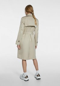 Gabardina beige con cintura ceñida, hombreras y abertura trasera. Fabricada en un tejido suave, cuenta con mangas largas y detalles de botones.