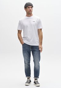 Camiseta blanca de algodón con logo impreso, combinada con jeans azules desgastados y zapatillas negras. Modelo posando contra un fondo liso.