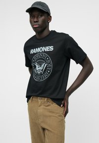 Tricou negru din bumbac cu inscripția "RAMONES" și un sigiliu grafic. Asortat cu pantaloni din stofa de corduroy bej, care prezintă linii verticale texturate.
