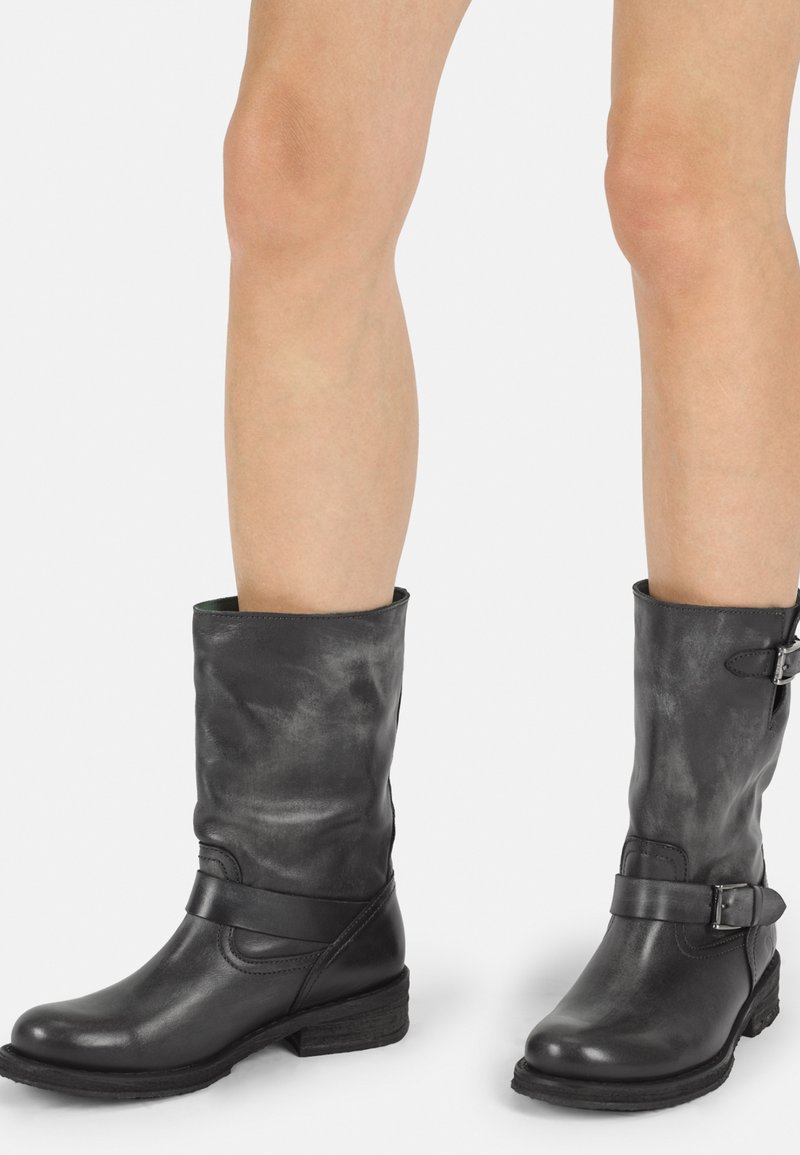 Botas de piel negra hasta la rodilla con puntera redondeada, correa en el tobillo y detalle de hebilla, que presentan una superficie ligeramente texturizada y un tacón bajo.