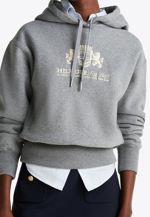 Osoba w szarej krótkiej bluzie z haftowanym herbem i napisem, nałożonej na białą koszulę oraz granatową spódnicę z złotym guzikiem.