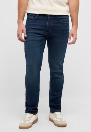 Mężczyzna ubrany w granatowe, dopasowane jeansy i białe trampki, stojący na jednolitym szarym tle.