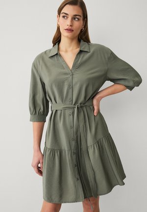 Kvinde iført en salviegrøn kjole med knapper, 3/4-ærmer og et bindebælte i taljen, stående med den ene hånd på hoften mod en ensfarvet baggrund.