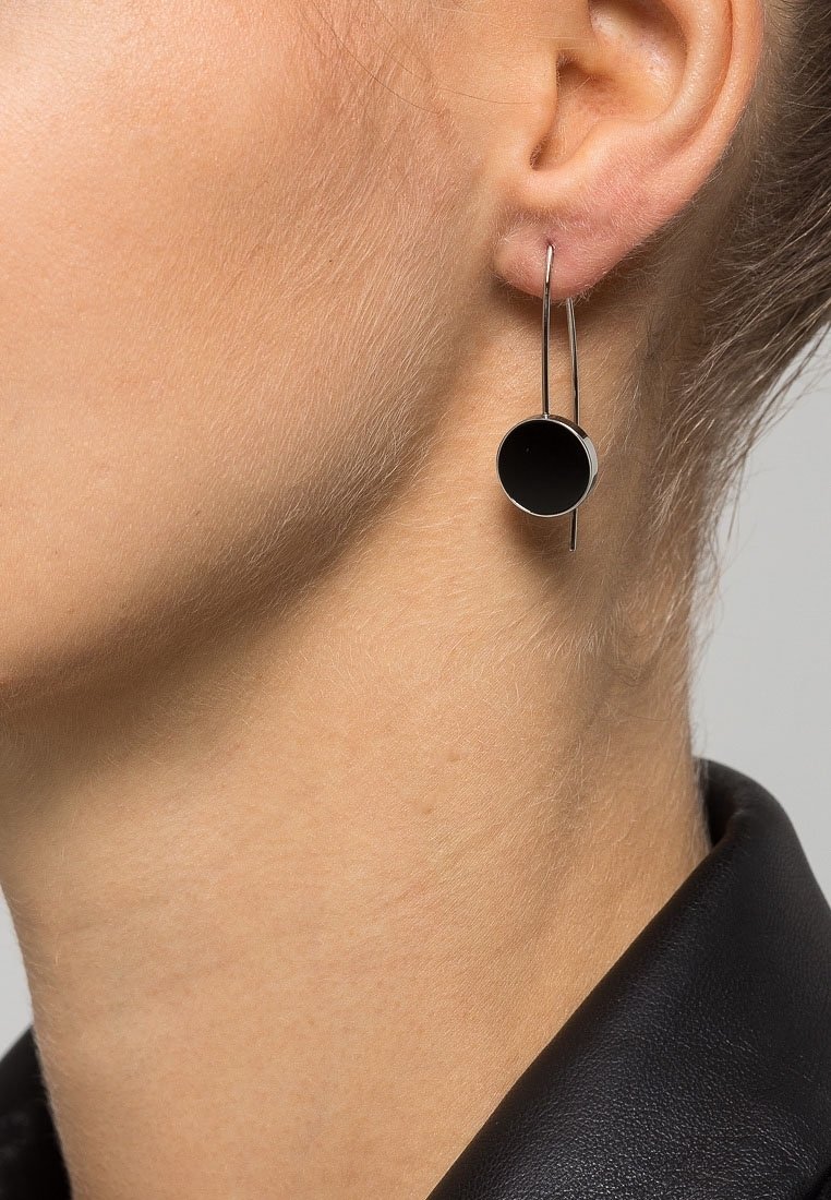 Oreille de femme portant une boucle d’oreille moderne en argent avec un pendentif circulaire noir suspendu à un fin fil courbé.