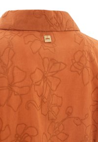 Camicia in tessuto color ruggine con motivo floreale ricamato, dotata di colletto e di una piccola etichetta dorata sul retro del collo.