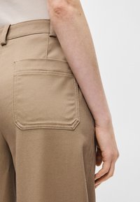 Beige kokvilnas bikses ar taisnu piegriezumu, ar redzamu aizmugurējo kabatu un teksturētu audumu. Jostasvieta atrodas tieši virs gurniem.