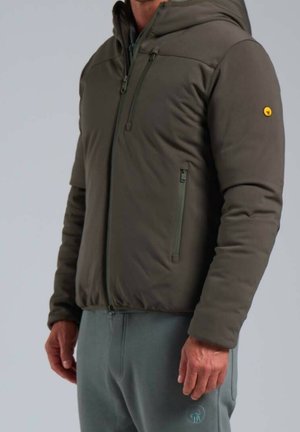 Mann trägt eine olivgrüne Kapuzenjacke mit Reißverschluss, Brust- und seitlichen Taschen mit Reißverschluss, kombiniert mit grauer Hose vor schlichtem Hintergrund.