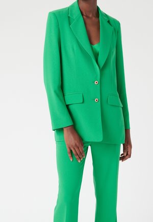 Personne portant un blazer vert vif ajusté avec des boutons décoratifs, un pantalon vert assorti et un haut vert en dessous.