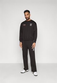 Fekete pamut keverék pulóver és nadrág szett. Fehér és piros hímzett logóval rendelkezik. A modellen fehér cipők vannak.