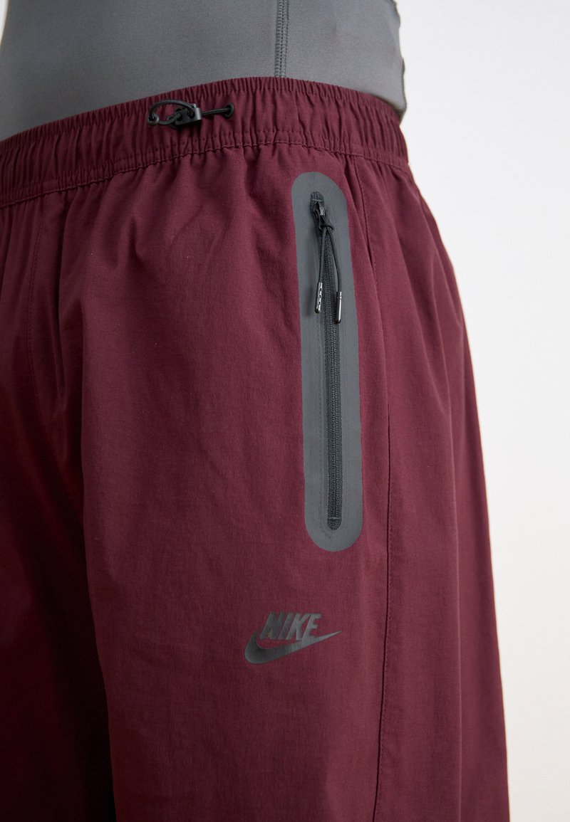 Pantalones cortos deportivos color burdeos con cintura elástica, un bolsillo lateral con cremallera y un acento gris. Presenta un sutil logo de Nike.