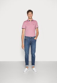 Camisa polo rosa claro con acentos de cuello azul marino, combinada con jeans ajustados azules y zapatillas blancas sin cordones, de pie contra un fondo liso.