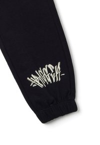 Schwarze Sweatpants-Bein mit elastischem Bund, versehen mit weißem Graffiti-ähnlichem Text im Knöchelbereich.