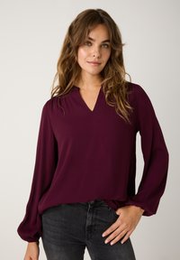 Burgunderfarbene Bluse mit langen Ärmeln, V-Ausschnitt und Puffärmeln, aus glattem Stoff, kombiniert mit dunklen Jeans.