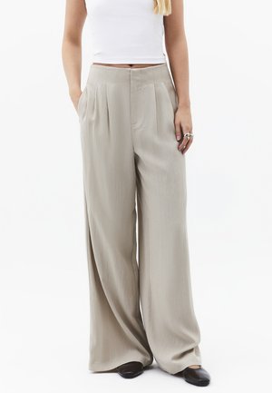 Femme portant un pantalon beige taille haute à jambes larges avec des plis sur le devant, associé à un haut court blanc et des chaussures plates noires.