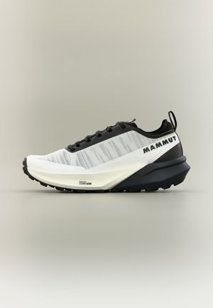 Zapatilla de trail running baja en colores blanco y negro con la marca Mammut y suela texturizada, vista de lado sobre un fondo neutro.