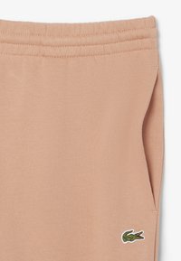 Pantaloni della tuta color pesca con vita elastica, tasca laterale, dettagli a coste e un piccolo logo di coccodrillo ricamato in verde.