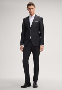 Schwarzer Anzug mit maßgeschneidetem Design, bestehend aus einer einreihigen Jacke, mit Reverskragen und schmal geschnittenen Hosen, kombiniert mit einem weißen Hemd.