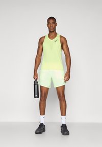 Canottiera sportiva gialla neon e pantaloni corti realizzati in materiale leggero, con un design texturizzato e dettagli del logo. Bottiglia d'acqua in mano.