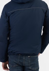 Homme portant une veste bleu foncé et un jean bleu, vu de dos, les mains près des poches.