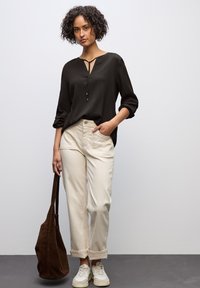 Blusa nera e testurizzata con scollatura a foro chiave, abbinata a pantaloni beigi chiari con risvolto. Il modello porta una borsa in suede marrone e indossa scarpe da ginnastica bianche.