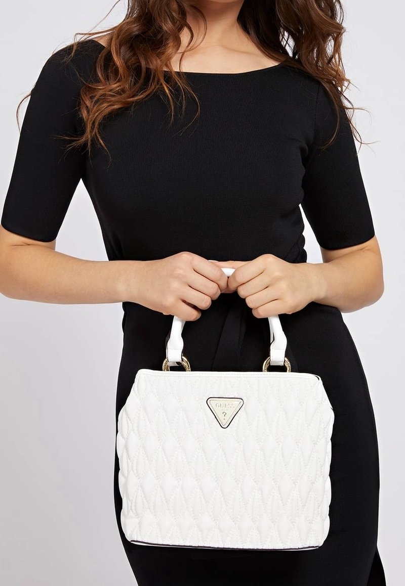 Femme en robe noire tenant un sac à main matelassé blanc avec des ferrures dorées et un logo triangulaire à l’avant.