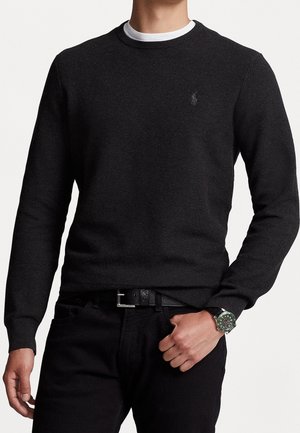 Maglione nero a maglia con colletto rotondo, maniche lunghe e un piccolo logo ricamato sul petto, indossato sopra dei pantaloni neri e un orologio.