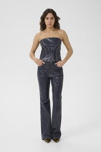 Mujer de pie con las manos en los bolsillos, vistiendo un top corset oscuro de mezclilla sin tirantes y jeans acampanados a juego, frente a un fondo claro y sencillo.