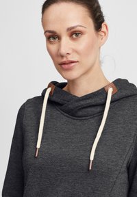 Mørk grå hettegenser med cowl-hals, hvite snorer med metalltips, og brune lærdetaljer på halsen. Myk teksturert stoff.