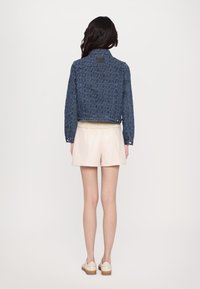 Dunkelblaue Jeansjacke mit geometrischem Muster, kurz geschnitten und mit geknöpften Manschetten. Kombiniert mit hellbeigen Shorts, Modell von hinten gewendet.