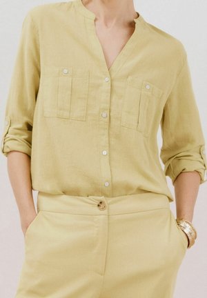 Frau trägt ein hellgelbes, langärmliges Hemd mit hochgekrempelten Ärmeln und passende hoch taillierte Hose, Hände in den Taschen und ein goldenes Armband.