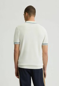 Weißes Strick-Polohemd mit gerippter Textur, das helle blaue Akzentstreifen am Kragen und an den Ärmeln aufweist. Getragen mit marineblauen Hosen.