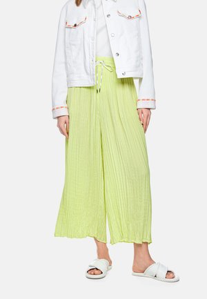 Weiße Jeansjacke mit farbiger Einfassung, getragen über einem weißen Top, kombiniert mit hellgrünen, plissierten, weitgeschnittenen Hosen und weißen geflochtenen Pantoletten.