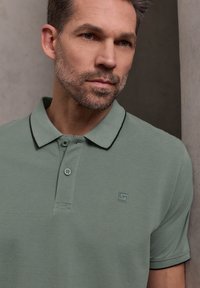 Homme à la barbe courte portant un polo vert sauge avec des bordures noires, appuyé contre un mur en béton gris, regardant sur le côté.