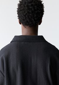 Indumento nero a maglia con un tessuto testurizzato, dotato di colletto e motivi a righe verticali, visto da dietro.