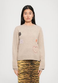 Maglione beige lavorato a maglia con dettagli ricamati, tra cui cuori e testo. Abbinato a una gonna dorata a strisce tigre.