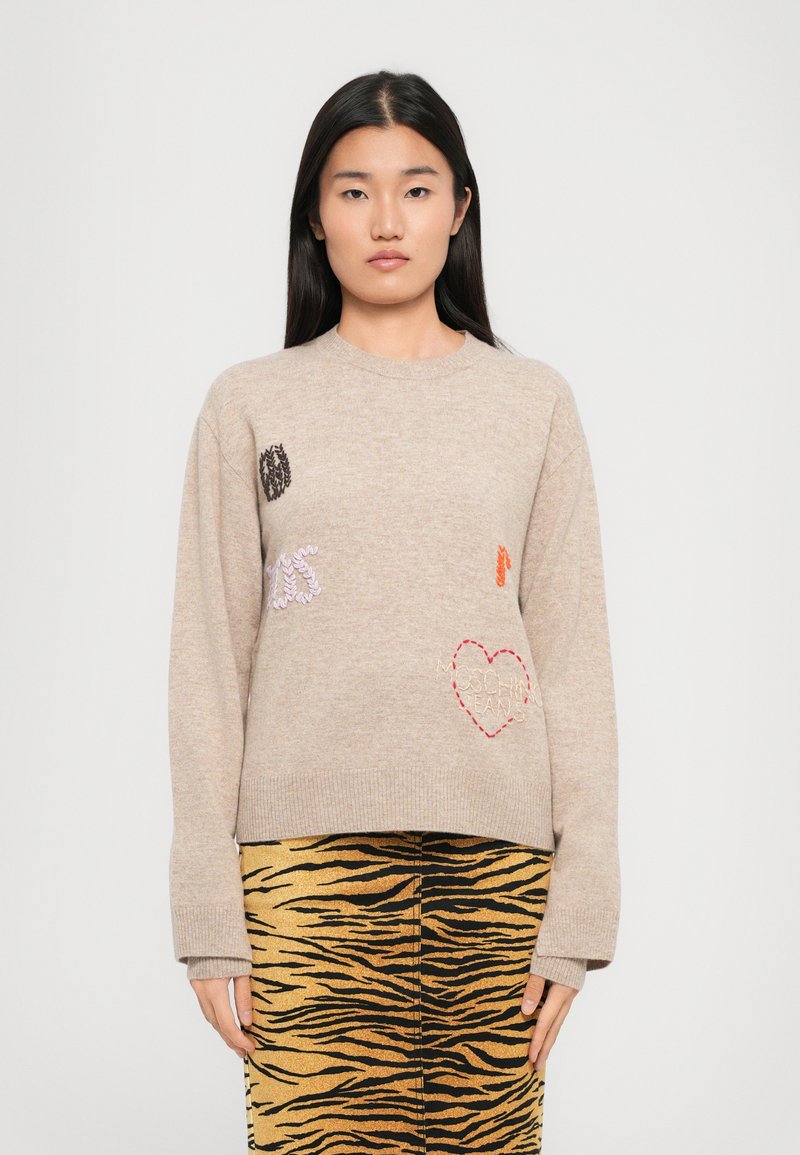 Maglione beige lavorato a maglia con dettagli ricamati, tra cui cuori e testo. Abbinato a una gonna dorata a strisce tigre.