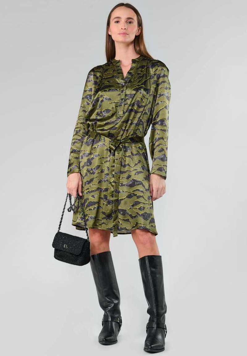 Olivgrünes Hemdkleid mit schwarzem Camouflage-Muster, Taillengürtel, langen Ärmeln und Knopfverschluss. Kombiniert mit schwarzen kniehohen Stiefeln und einer Handtasche.