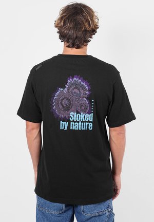 Personne portant un t-shirt noir avec un motif floral violet et bleu et le texte "Stoked by nature" au dos, porté avec un jean bleu.