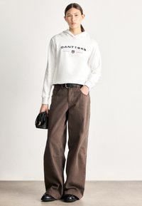 Hanorac alb cropped cu textul "GANT 1949", asortat cu pantaloni largi maro. Curea neagră și geantă mică neagră completează ținuta. Pantofi negri.