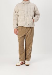 Vadderad beige jacka med hög krage, strukturerad tyg, med en framficka. Matchad med ljusbruna manchesterbyxor och vita skor med röda ränder.