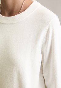 Witte gebreide trui met een ronde halslijn en geribbelde manchetten. Zachte textuur met een gladde afwerking, waarbij de schouder- en nekzone zichtbaar zijn.