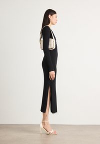 Black ribbed dress with side slit, paired with white strappy heels. A beige shoulder bag sits on the shoulder, adding contrast to the outfit.