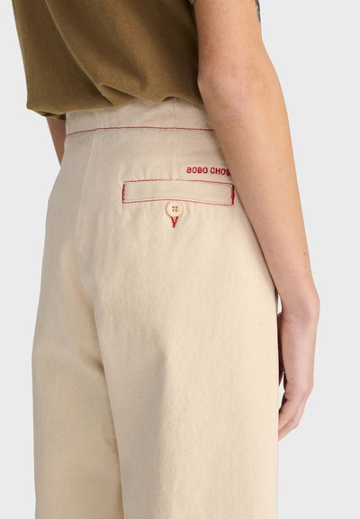 Gros plan d'une personne portant un pantalon beige avec des coutures rouges et une poche arrière boutonnée, haut marron partiellement visible.