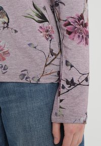 Långärmad överdel i mjukt lila tyg med blommönster och fågelmotiv. Denimjeans synliga längst ner med en avslappnad passform.